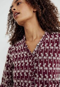 Blusa burdeos con bordado floral blanco, cuello con volantes y un diseño ajustado. Tela texturizada con patrones intrincados en todo su recorrido.