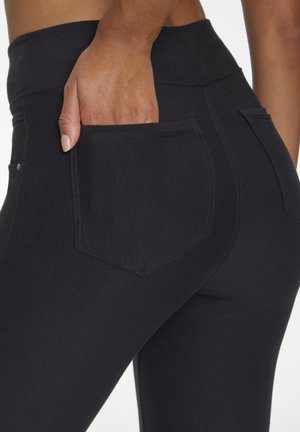 Eine Hand steckt in die Gesäßtasche einer schwarzen, eng anliegenden Hose, die von einer Person gezeigt wird, die von der Taille bis zur Mitte des Oberschenkels sichtbar ist.