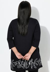 Schwarze Tunika mit Dreiviertelärmeln und weißer abstrakter Stickerei am Saum. Der Stoff wirkt glatt, mit einer lockeren Passform und Seitenschlitzen.