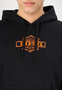 Svart hettegenser med frontlomme, med oransje grafisk logo-trykk. Materialet ser mykt ut, med justerbar snor i hetten.