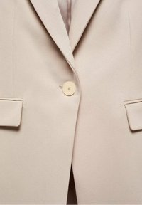 Blazer beige à texture lisse, fermeture à un bouton, et deux poches avant. Comprend un revers cranté et une coupe ajustée.