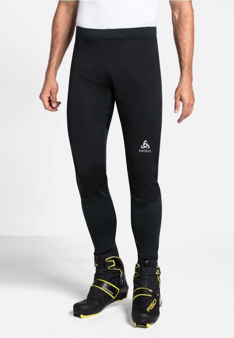 Mann in schwarzen Odlo-Sportleggings und schwarzen sowie gelben Fischer-Skischuhen steht vor weißem Hintergrund.