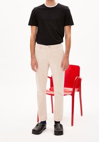 Černé tričko s krátkými rukávy a bílým límečkem, světle béžové kalhoty a černé dřeváky. V pozadí červená židle.