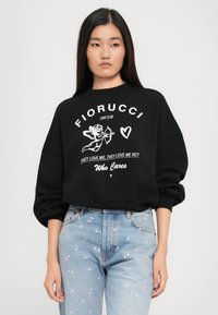 Jovem mulher a usar um hooded preto com um gráfico de cupido e texto, combinado com jeans azul claro com pequenos padrões de flores rosas.