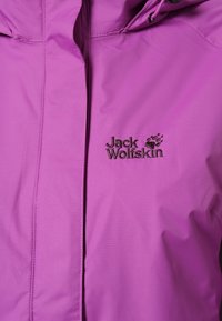Chaqueta impermeable morada con cierre frontal de cremallera, capucha ajustable y logo bordado de "Jack Wolfskin". Textura suave y material ligero.