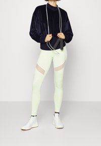 Sweat-shirt en velours bleu marine associé à des leggings vert clair avec des découpes en mesh. Le mannequin tient une corde à sauter ; des chaussures de sport blanches complètent la tenue.
