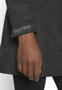 Črna jakna Helly Tech s bližnjim posnetkom roke, ki drži manšeto, z oznako logotipa "Helly Tech® Protection" v svetlobno modri barvi.