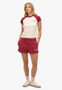 Camiseta de algodón color burdeos y crema con mangas raglán, combinada con shorts burdeos que llevan impreso "SUPERDRY NYC". Zapatillas de color claro completan el look.