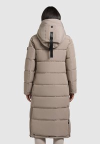 Beige Steppjacke mit langem Schnitt, horizontalen Nähten, einer großen Kapuze und einem schwarzen Emblem auf der Rückseite. Mit Reißverschlusstaschen ausgestattet.