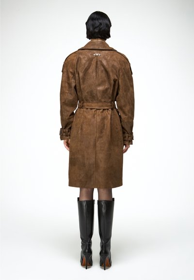 Osoba s krátkymi tmavými vlasmi, oblečená v hnedom semišovom trenčkote so stužkou v páse a čiernych kozačkách s podpätkom po kolená, stojaca chrbtom k nám na bielom pozadí.