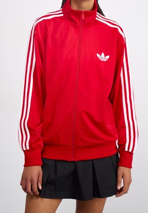 Personne portant une veste rouge zippée avec des rayures blanches sur les manches et une jupe plissée noire sur un fond uni.
