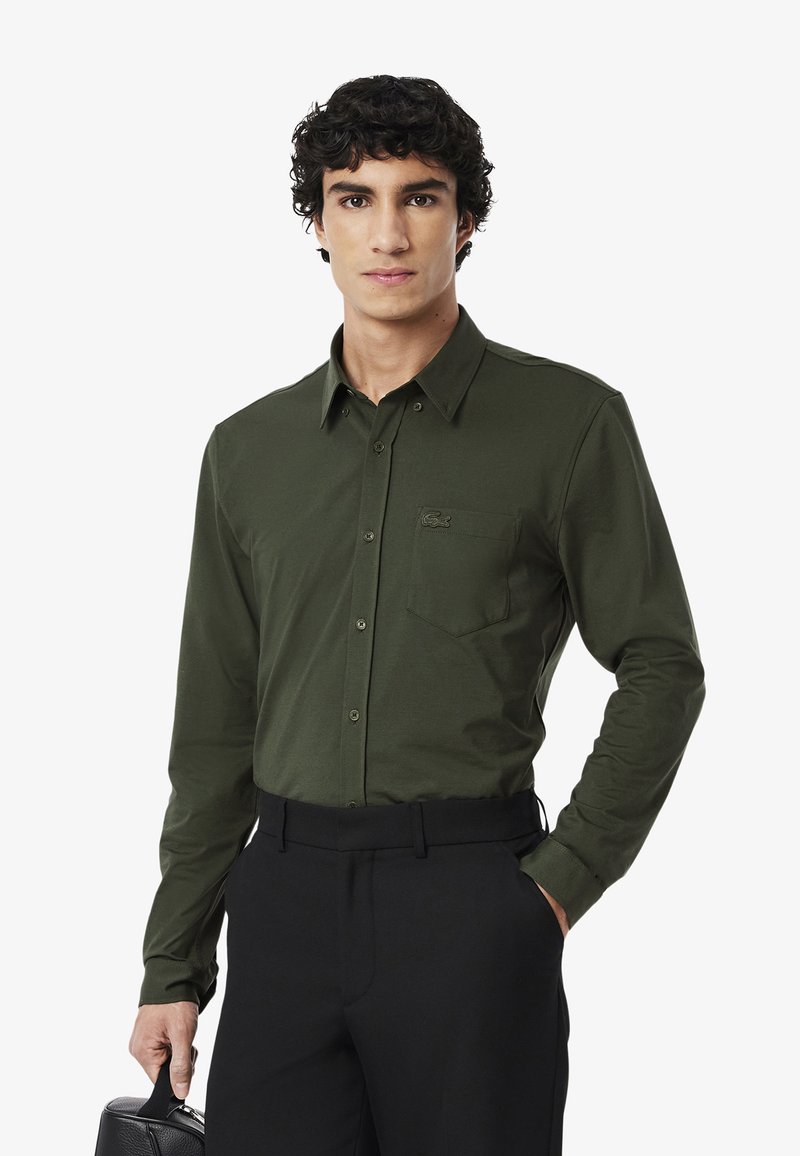 Camicia a maniche lunghe verde scuro con colletto button-down, tasca frontale e logo sottile; abbinata a pantaloni neri e con una borsa nera.