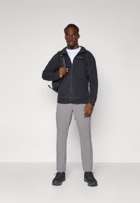 Chaqueta de forro polar negra con cremallera, pantalones grises y botas de senderismo negras. El modelo sostiene una mochila, de pie contra un fondo blanco liso.