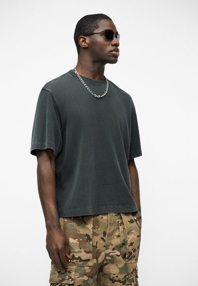 Άνδρας που φοράει μαύρα γυαλιά ηλίου, γκρι σκούρο φαρδύ μπλουζάκι, ασημένιο αλυσίδα λαιμού και παντελόνι καμουφλάζ cargo σε λευκό φόντο.