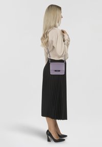 Fioletowa skórzana torebka crossbody o sztywnej konstrukcji, z srebrnym okuciem, noszona z długą czarną plisowaną spódnicą i czarnymi szpilkami.