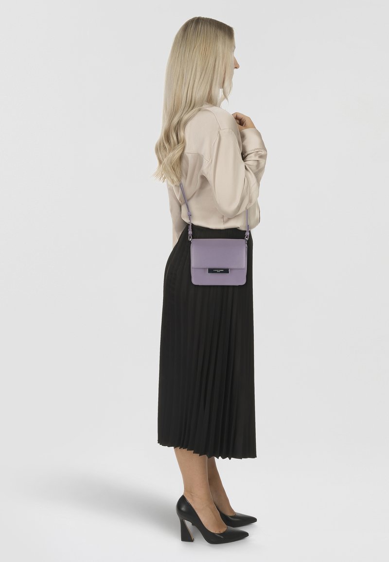 Fioletowa skórzana torebka crossbody o sztywnej konstrukcji, z srebrnym okuciem, noszona z długą czarną plisowaną spódnicą i czarnymi szpilkami.