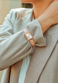Reloj rectangular de acero inoxidable con esfera plateada, correa de metal dorado y manga de chaqueta con patrón de espiga texturizado.