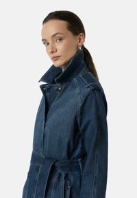 Donkerblauwe denim trenchcoat met een hoge kraag, knoopsluiting, ceintuur in de taille en dubbele zijzakken, voorzien van een gestructureerde stof en een getailleerde pasvorm.
