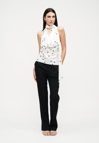 Mujer con cabello largo y oscuro, que lleva una blusa de tirantes blanca con lunares y pantalones negros, de pie con una mano en el bolsillo.