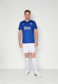 Modrý fotbalový dres s vlnkovým vzorem, bílým logem "Stake.com", v kombinaci s bílými šortkami a ponožkami, s černo-bílými botami.