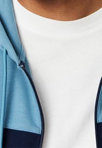 Veste à capuche en tissu bleu clair et bleu marine avec une fermeture éclair à l'avant, portée sur un t-shirt blanc à encolure ronde. Texture lisse, design décontracté.
