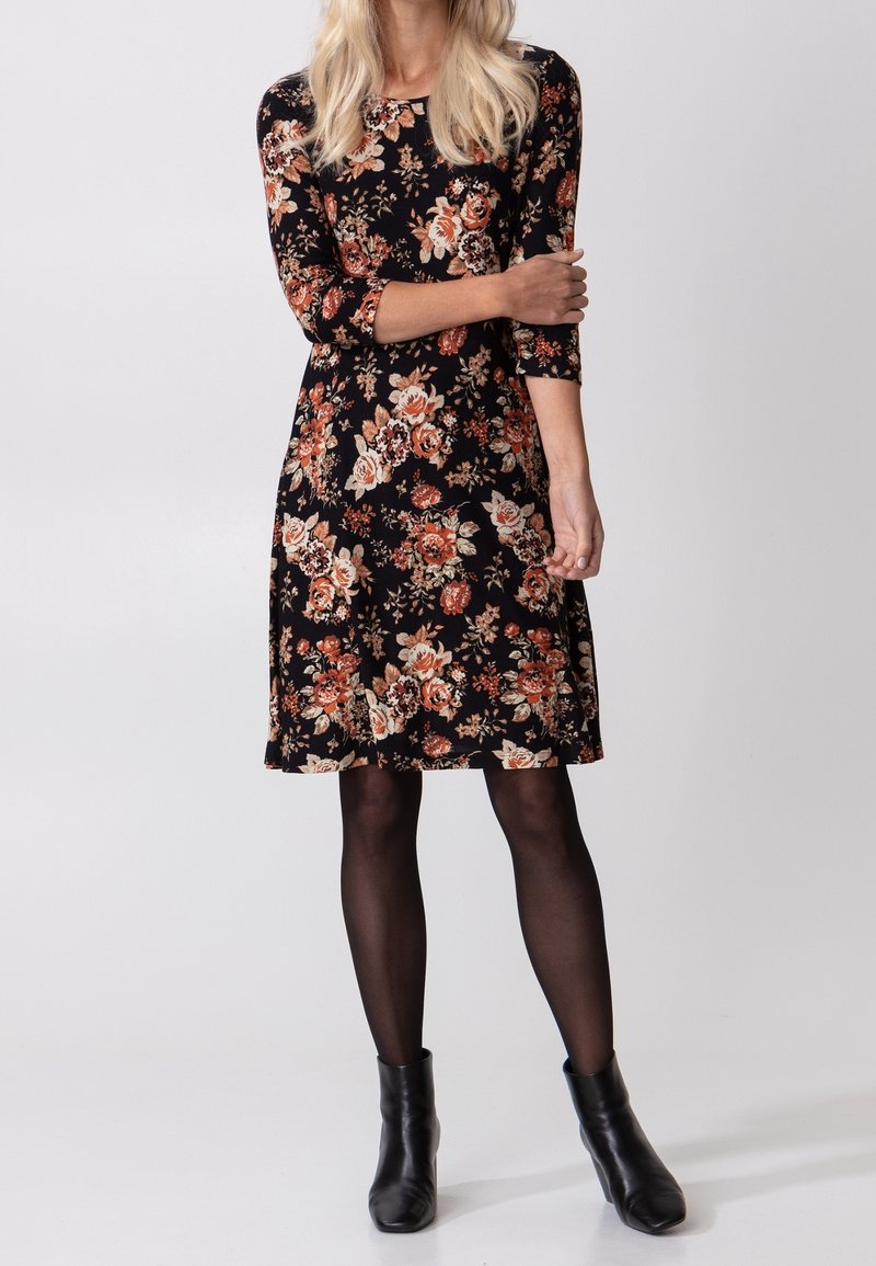 Femme portant une robe noire à fleurs mi-longue avec des manches trois quarts, des collants noirs et des bottines noires, debout avec un bras croisé.