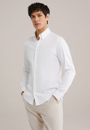 Camicia bianca con bottoni in tessuto leggero e finitura morbida, con colletto classico e maniche lunghe. Indossata con pantaloni beige.