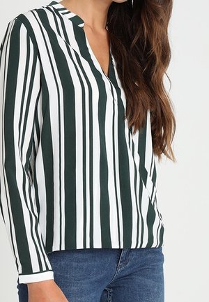 Kvinde iført en langærmet hvid bluse med mørkegrønne lodrette striber og blå jeans, stående mod en ensfarvet baggrund.