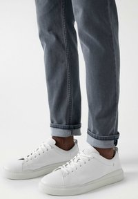Zapatillas de cuero blancas con acabado texturizado, puntera redonda y diseño de cordones. Llevadas con jeans de denim gris remangados, que presentan un aspecto limpio y ajustado.