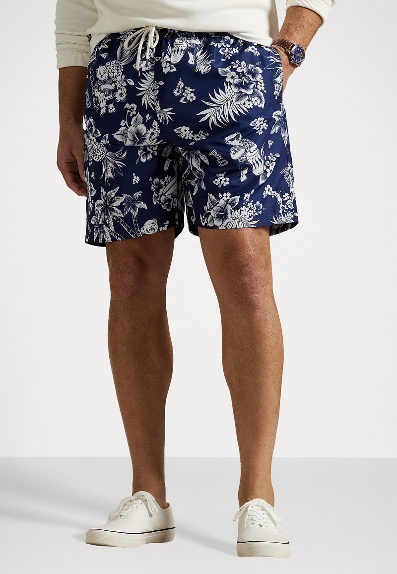 Jūras zila peldbikses ar baltu tropisku ziedu rakstu, elastīga josta ar auklu, un sānu kabatām. Valkātas baltas sportsnes.