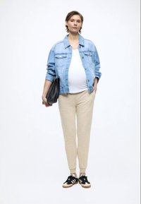 Casaco de ganga, top branco, calças de fato de treino bege e sapatilhas pretas. Modelo segura uma mala preta, com foco no design confortável e casual.