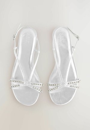 Sandales argentées à brides, présentant une finition brillante, des brides transparentes et des embellissements en strass sur la zone des orteils et la bride de cheville.