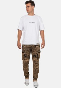 Maglietta bianca a maniche corte con logo nero. Pantaloni cargo marroni stone-washed con molte tasche e dettagli grafici neri. Sneakers bianche.