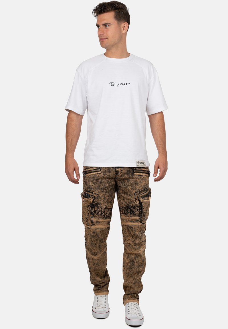 Maglietta bianca a maniche corte con logo nero. Pantaloni cargo marroni stone-washed con molte tasche e dettagli grafici neri. Sneakers bianche.