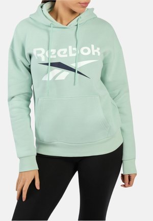 Hellgrüner Reebok-Hoodie, getragen von einer Person mit schwarzen Leggings, zeigt das Frontlogo und die Kängurutasche, Hände ruhen nahe an den Hüften.