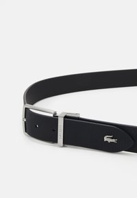 Sort læderbælte med perforeret tekstur, sølvfarvet metalspænde og Lacoste-branding. Har et krokodillelogo på remmen.