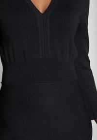 Svart stickad klänning med V-ringning och långa ärmar, med ett texturerat ribbat mönster vid midjan och slät tyg nedanför.