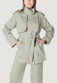 Cappotto trench verde chiaro con cintura, colletto alto e tasche laterali. Presenta una chiusura a bottoni e un design stratificato e strutturato.