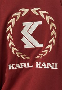Logo "K" ricamato in bianco, circondato da una corona di alloro beige sopra il testo ricamato in bianco "KARL KANI" su tessuto rosso.