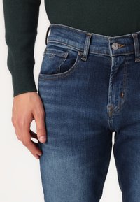 Blugi din denim de culoare albastru închis, cu o croială slim, având un design clasic cu cinci buzunare, ușor decolorati și detalii de cusătură în contrast.