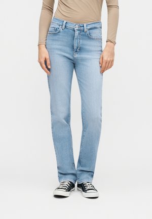 Άτομο που φοράει ανοιχτό μπλε τζιν με ίσια ποδιά, μπεζ μπλούζα με μακριά μανίκια και μαύρα-λευκά αθλητικά παπούτσια με κορδόνια, στέκεται μπροστά σε λευκό φόντο.