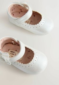 Baltas ādas zīdaišu kurpes ar viļņotām malām, ar siksniņu un loku. Mīksta rozā iekšpuse ar ziedu rakstu. Izmērs 0-3 mēneši.