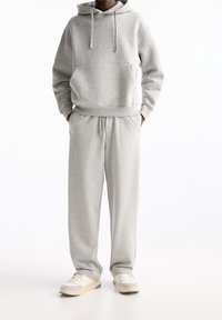 Grauer Hoodie mit einer frontalen Tasche und Kapuze mit Kordelzug, kombiniert mit passender grauer Sweatpants. Die Sneakers haben ein weißes Obermaterial und beige Akzente.