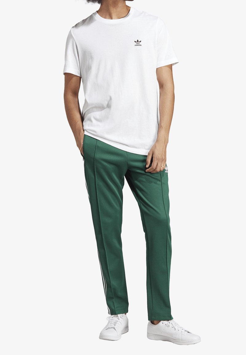 Vit bomull t-shirt med en liten svart logotyp, i kombination med gröna träningsbyxor med vita sidostreck och vita sneakers.