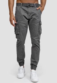 Pantalon cargo gris avec une taille ceinturée, des poignets élastiqués et plusieurs poches latérales, confectionné dans un tissu doux et durable. Porté avec des baskets blanches.