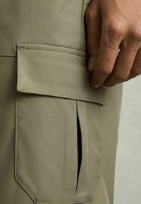 Pantaloni cargo khaki cu o buzunare cu clapetă și o deschidere laterală, realizați dintr-un țesătură netedă și durabilă. Designul are linii simple și clare.