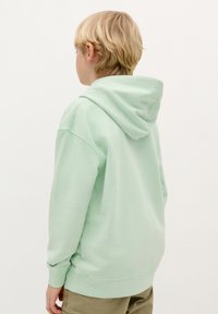 Felpa verde menta con tasca a manica sul davanti, vestibilità over, cappuccio con cordino e polsini a coste. Indossata sopra pantaloni beige chiaro.