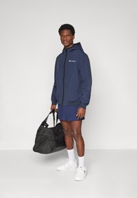 Navy windbreaker with zipper, logo on chest, elastic cuffs, worn over blue shorts; holding a black duffel bag; white sneakers and socks.