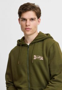 Oliven grønn zip-up hettegenser laget av mykt stoff, med kengurulomme og brodert "Be Everything and More" design i rosa.