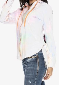 Veste zippée irisée blanche avec des cordons orange, présentant un ourlet asymétrique et un tissu texturé, associée à un jean bleu délavé ajusté.